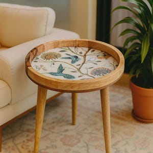 Puede incluir: Una mesa auxiliar redonda de madera con un diseño decorativo de flores y pájaros en la mesa. La mesa tiene tres patas anguladas y un borde elevado. La mesa presenta un fondo crema con pájaros azules, flores y ramas.
