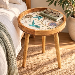 May include: A round wooden side table with three tapered legs and a tray-style top. The tabletop features a cream-coloured surface decorated with a botanical print of teal hummingbirds, brown branches, and multi-coloured flowers, topped with a book.