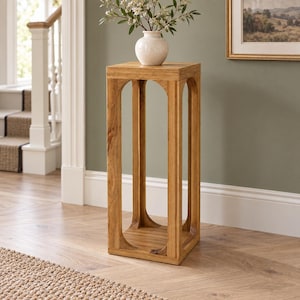 Puede incluir: Un pedestal cuadrado de madera alto con un acabado de veta de madera marrón claro natural. La estructura cuenta con cuatro patas verticales conectadas por recortes arqueados en la parte superior e inferior. Un jarrón de cerámica blanca con flores blancas descansa encima.