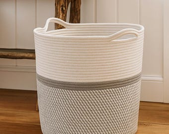 May include: A cylindrical storage basket constructed from coiled rope. The upper portion is solid off-white, while the lower section features a textured, woven pattern in alternating gray and white tones. It includes two integrated rope handles.