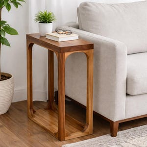 May include: A wooden side table with a rectangular top and arched legs. The table is made of light and dark brown wood. A small potted plant, a book, and glasses rest on the table's surface. The table is next to a light-coloured sofa.