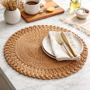 Puede incluir: Un mantel individual redondo y tejido en tonos naturales con un borde festoneado. Un plato blanco, una servilleta doblada y cubiertos dorados están encima. El mantel individual está sobre una superficie de mármol blanco, con una taza de café y flores secas.