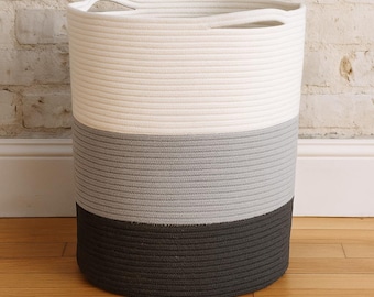 May include: A cylindrical storage basket with a three-tone design. The top section is off-white, the middle is gray, and the bottom is dark gray. The basket has two integrated handles and is made of a woven rope material.