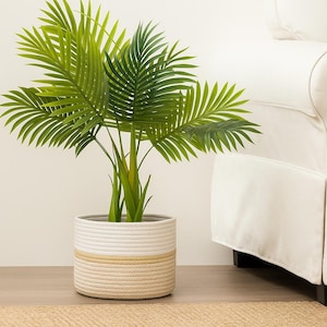May include: A potted artificial palm plant with vibrant green fronds. The plant is in a round, woven basket with a white top, a yellow stripe, and a beige bottom. The plant is placed on a wooden floor.