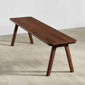 Könnte beinhalten: Eine dunkelbraune Holzbank mit einer glatten, rechteckigen Sitzfläche mit abgerundeten Ecken und vier angewinkelten, konisch zulaufenden Beinen. Die Holzmaserung ist auf der Sitzfläche sichtbar und zeigt ein natürliches, tiefes Finish.