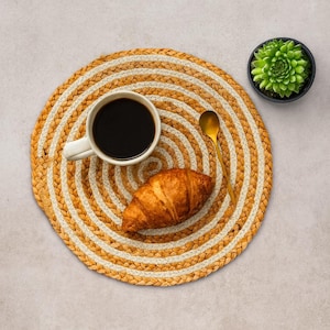 Op de afbeelding: Een ronde, geweven placemat met afwisselende beige en witte concentrische cirkels. Een witte mok gevuld met zwarte koffie, een gouden lepel en een goudbruine croissant liggen op de placemat. Een kleine vetplant in pot staat in de rechterbovenhoek.