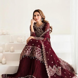 Op de afbeelding: Een bordeauxrode, geborduurde formele jurk met een bijpassende dupatta. De jurk heeft ingewikkelde gouden borduursels op het lijfje, de mouwen en de zoom. Het model zit op witte trappen, met kaarsen op de achtergrond.