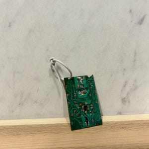 May include: A small, rectangular green circuit board with copper traces and electronic components. A white wire is attached to the top of the board. The board is resting on a wooden surface against a white and gray background.