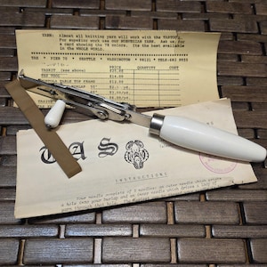 May include: A vintage TAS tool with a white handle and silver metal components, resting on a wooden surface. Accompanying it are instruction sheets and a price list for various items, including burlap. The text on the paper reads "TAS TOOL".
