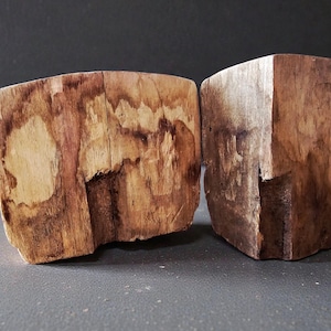 May include: Two rustic wooden blocks with natural wood grain patterns. The blocks are rectangular with a rough-hewn appearance, showcasing variations in color from light tan to dark brown. The wood has visible knots and imperfections.