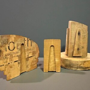 May include: Three wooden sculptures of varying sizes and shapes. The largest has text fragments and a cutout. The middle one is rectangular with a triangular cutout. The third is on a round base.