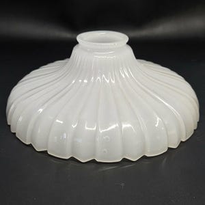 May include: A white milk glass lamp shade featuring a wide, shallow, bowl-like shape with a distinct fluted or ribbed pattern radiating from the center opening toward the scalloped edge. The glass has a semi-translucent, smooth, glossy finish.