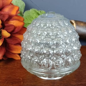 May include: Clear glass light shade with a textured, bubble-like surface. The shade is dome-shaped and sits on a wooden surface, with a decorative sunflower and foliage in the background.