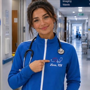 May include: A blue zip-up jacket with white trim and the text "Lara, RN" with a heart and pulse design. A stethoscope hangs around the neck. The jacket is worn in a hospital setting.
