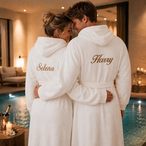 May include: Two white hooded bathrobes made of plush fabric, each featuring long sleeves and a tie belt. The left robe is embroidered with the name Selena in gold script, and the right robe is embroidered with the name Harry in gold script.