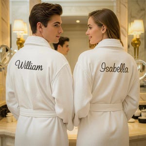 May include: Two white terry cloth bathrobes with the names "William" and "Isabella" embroidered on the back. The robes have a shawl collar and a matching belt. The couple is standing in a bathroom with a mirror and lamps.