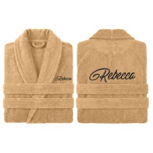 May include: Two views of a beige terry cloth robe with a shawl collar and a waist tie. The name "Rebecca" is embroidered in a cursive font on the front and back of the robe. The robe is made of a soft, absorbent material.