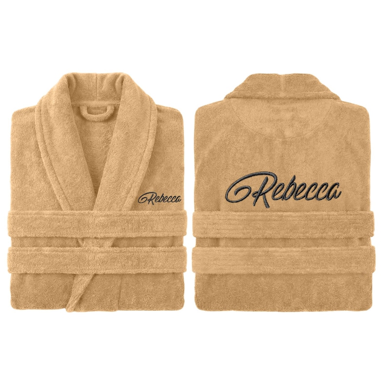May include: Two views of a beige terry cloth robe with a shawl collar and a waist tie. The name "Rebecca" is embroidered in a cursive font on the front and back of the robe. The robe is made of a soft, absorbent material.