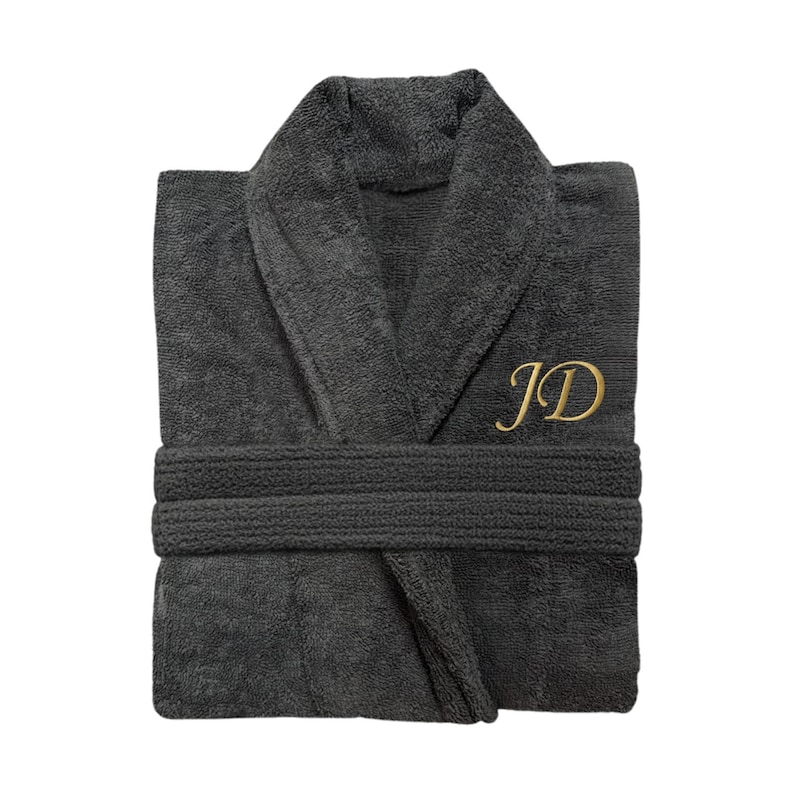 Puede incluir: Bata de ba&ntilde;o de rizo gris oscuro con cuello chal y cintur&oacute;n a juego. La bata presenta las iniciales doradas "JD" bordadas en el pecho. La bata est&aacute; cuidadosamente doblada, mostrando su textura afelpada y dise&ntilde;o.