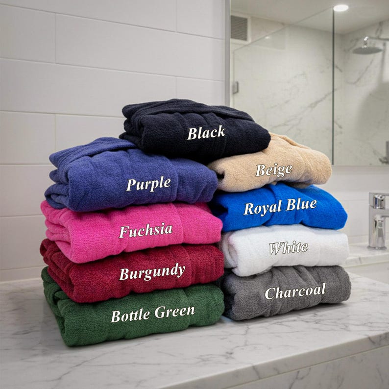 May include: A stack of folded bathrobes in various colors, including black, purple, fuchsia, burgundy, bottle green, beige, royal blue, white, and charcoal. Each robe has a label indicating its color. The robes are neatly arranged on a marble countertop.