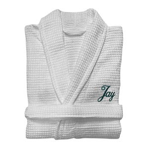 May include: White waffle-weave dressing gown with a shawl collar and a matching belt. The name "Jay" is embroidered in green script on the front. The gown is folded, highlighting its textured fabric and design.