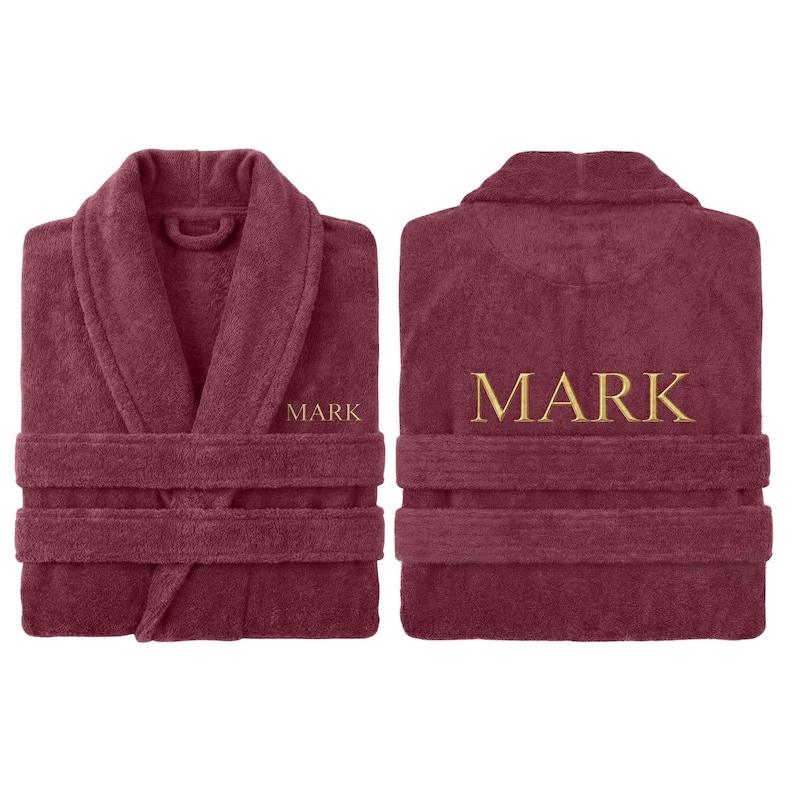 May include: A burgundy terry cloth robe with a shawl collar and belt loops. The name "MARK" is embroidered in gold thread on the front and back of the robe. The robe is displayed from the front and back.