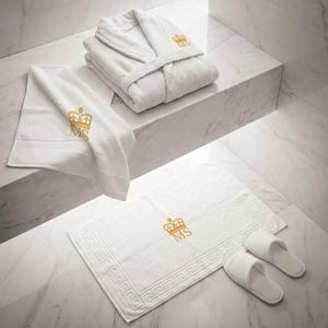 May include: A luxurious white bathroom set featuring a plush robe, a hand towel, a bath mat, and a pair of slippers. Each item is embroidered with a gold crown and the initials "MS". The bath mat has a Greek key border.