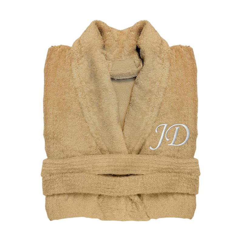 Puede incluir: Una bata de felpa plegada, de color beige, con cuello chal y cintur&oacute;n. La bata presenta las iniciales "JD" bordadas en blanco en el pecho. Fabricada con un material suave y absorbente, ideal para despu&eacute;s de la ducha.