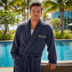 May include: A navy blue terry cloth robe with the name "SEAN" embroidered in gold. The robe has a shawl collar, a waist tie, and pockets. The person is standing near a pool with palm trees in the background.