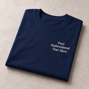 May include: A navy blue crew neck t-shirt featuring white embroidered text on the chest that reads Your Embroidered Text Here. The garment is folded neatly, showcasing the smooth fabric texture and the clean, centred placement of the embroidery.