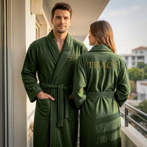 May include: Two people wearing matching dark green bathrobes with gold embroidered names. The robes have shawl collars, pockets, and waist ties. The person on the left has the name "SE" embroidered on the chest, and the person on the right has "TRACY" embroidered on the back.