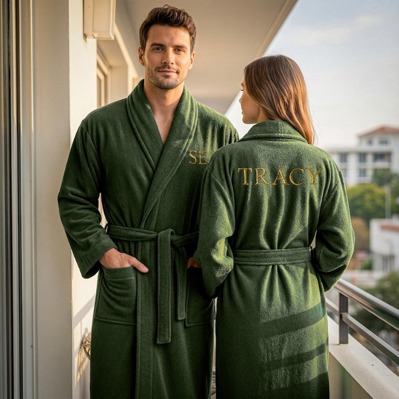 May include: Two people wearing matching dark green bathrobes with gold embroidered names. The robes have shawl collars, pockets, and waist ties. The person on the left has the name "SE" embroidered on the chest, and the person on the right has "TRACY" embroidered on the back.