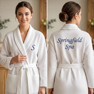 May include: A white waffle-knit bathrobe featuring a shawl collar, a tie belt, and two front pockets. The left chest is embroidered with a navy blue letter S, while the back displays the navy blue embroidered text Springfield Spa.