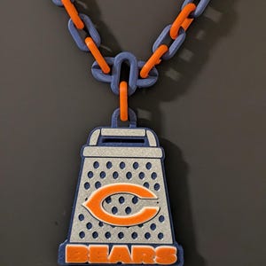 May include: A necklace featuring a large pendant shaped like a cheese grater with a silver glitter finish. The pendant displays an orange Chicago Bears 'C' logo and the word 'BEARS' in orange letters. The chain consists of alternating blue and orange links.