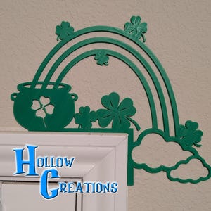 May include: A green St. Patrick's Day decoration featuring a rainbow, pot of gold, and shamrocks. The rainbow arches over a pot of gold with a shamrock cutout. The design includes shamrocks and a cloud. The text "Hollow Creations" is at the bottom.