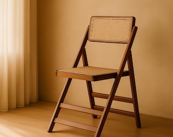 Pode incluir: Cadeira dobrável de madeira com assento e encosto em rattan tecido. A cadeira tem um acabamento castanho quente e um design simples e clássico. Está colocada contra uma parede de cor neutra e um chão de madeira.