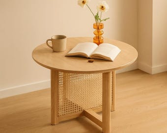 Pode incluir: Uma mesa lateral redonda de madeira com um painel de cana tecida e três pernas. Uma caneca bege, um livro aberto e um pequeno vaso laranja com flores brancas estão sobre a mesa. A mesa tem cerca de 61 cm de diâmetro.