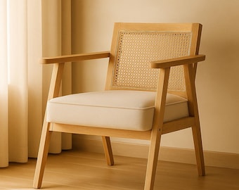 Pode incluir: Poltrona de madeira clara com encosto e apoios de braços em cana tecida. A almofada do assento é de cor creme claro. A cadeira está colocada sobre um fundo neutro, sugerindo uma estética de design minimalista ou escandinavo.