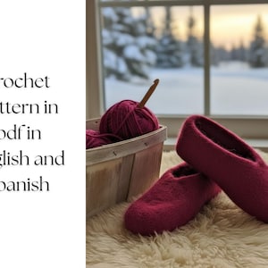 May include: A pair of deep pink felted slippers rests on a fluffy white rug, next to a basket holding yarn and a crochet hook. The text on the left says "crochet pattern in pdf in english and spanish". A wintery scene is visible through the window.