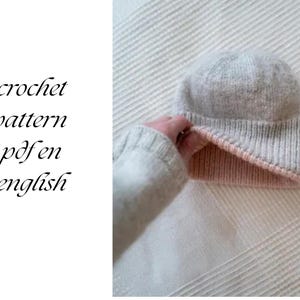 Könnte beinhalten: Hellgraue Strickmütze mit rosa Krempe. Der Text "crochet pattern pdf en english" befindet sich links. Die Mütze wird von einer Person gehalten. Die Mütze wirkt weich und warm, geeignet für kaltes Wetter.