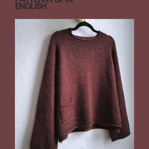 Può includere: Un maglione bordeaux oversize con una singola tasca, appeso a una gruccia nera contro un muro bianco. Il testo "CROCHET PATTERN PDF IN ENGLISH" è visibile nell'immagine.