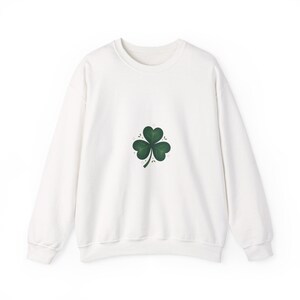 May include: A white crew neck sweatshirt with a green shamrock motif. The shamrock, centrally placed, has three heart-shaped leaves with fine detailing. The sweatshirt has long sleeves and a classic, casual design, suitable for St. Patrick's Day.