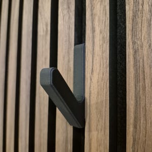 Könnte beinhalten: Ein dunkelgrauer Metallhaken an einer Holzlattenwand. Der Haken ist abgewinkelt und ragt nach außen, zum Aufhängen von Gegenständen. Das Holz hat ein natürliches Maserungsmuster mit dunklen vertikalen Streifen.