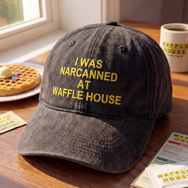 May include: A dark gray baseball cap with the text "I WAS NARCANNED AT WAFFLE HOUSE" embroidered in yellow. The cap is sitting on a wooden surface, with a waffle and a coffee mug in the background.