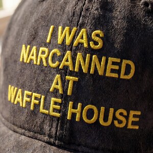 May include: A dark gray baseball cap with the embroidered text "I WAS NARCANNED AT WAFFLE HOUSE" in yellow thread. The cap is made of a textured fabric and has a curved brim.