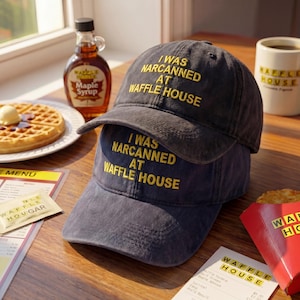 May include: Two dark gray baseball caps stacked on top of each other, each with the yellow text "I WAS NARCANNED AT WAFFLE HOUSE." The caps are on a wooden surface, with a waffle, syrup, and a coffee mug in the background. The mug says "WAFFLE HOUSE".