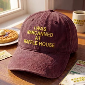 May include: A maroon baseball cap with the text "I WAS NARCANNED AT WAFFLE HOUSE" embroidered in yellow. The cap is sitting on a wooden surface near a window, with a waffle and a coffee mug in the background.