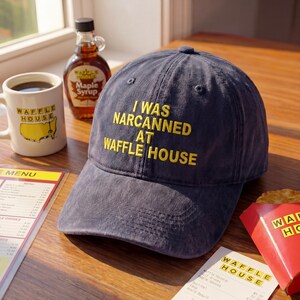 May include: A navy blue baseball cap with the text "I WAS NARCANNED AT WAFFLE HOUSE" in yellow. The image also shows a Waffle House mug, a bottle of maple syrup, and a menu.