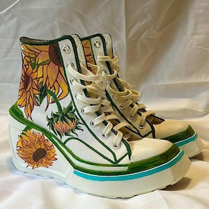 Könnte beinhalten: Weiße High-Top-Sneaker mit Keilabsatz, handbemalt mit Sonnenblumen. Das Design zeigt gelbe, grüne und braune florale Elemente, mit grünen Zierleisten und einem blauen Akzent auf der Sohle. Die Sneaker haben weiße Schnürsenkel.