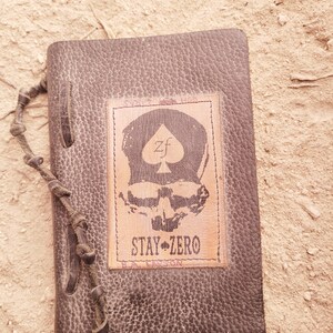 May include: A dark brown leather journal with a textured surface. The cover features a rectangular patch with a skull design and the text "STAY ZERO". A knotted cord closure is visible on the left side.